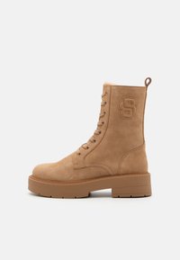 Kamelpruun suède pahklajalanikud tugevate taldadega, nöörimise disainiga ja logo aksessuaari korral küljel. Omab pehme sisepinda ja tõmbluku silti.