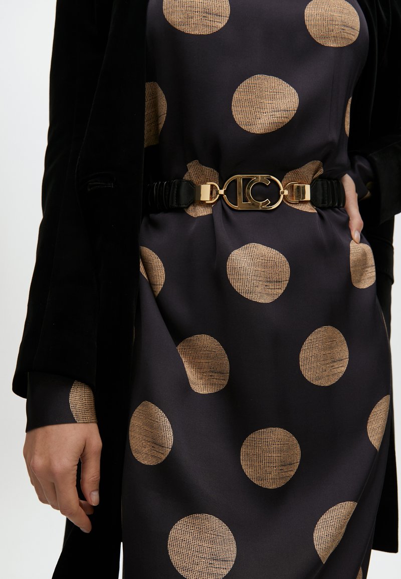 Schwarzes Kleid mit großen beigefarbenen Punkten, ausgestattet mit einem schwarzen Satinhochzug mit einer goldenen Metallschnalle und Logodetails. Samtstoffakzente an den Ärmeln.