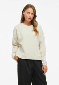 Felpa beige chiaro con collo a giro e ricamo "TRUTH" sulla parte anteriore. Presenta spalle cadenti e polsini a coste, abbinata a pantaloni neri.