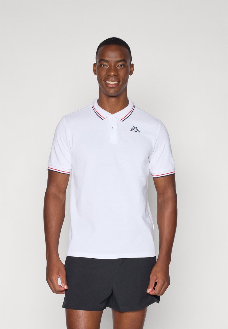 Hvit polo skjorte med stripete krage, korte ermer og en liten logo på brystet. Komplementert av svarte shorts.