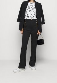 Czarna marynarka na białej bluzce z wzorem łańcucha, w zestawieniu z czarnymi spodniami o wysokim stanie z złotymi guzikami oraz białymi trampkami.