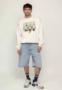 Young man wearing a cream sweatshirt with a running club graphic, light denim shorts, striped socks, and white sneakers, standing against plain background.