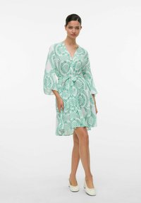 Grön mönstrad klänning med V-ringning, 3/4 ärmar med klockform, knuten vid midjan, med ett lätt tyg och en svängd fåll. Bärs med vita skor.