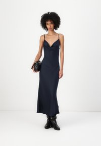 Ναυτικό σατέν φόρεμα με μπούστο σε σχήμα κουβέντας, λεπτές τιράντες και εφαρμοστή σιλουέτα, συνδυασμένο με μια μαύρη τσάντα και μαύρες μπότες μέχρι το γόνατο.