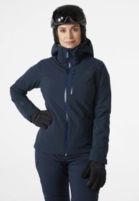 Marineblaue Skijacke mit Kapuze, ausgestattet mit einem Frontreißverschluss, Reißverschlusstaschen und schwarzen Handschuhen. Das Material scheint glatt und wetterbeständig zu sein.
