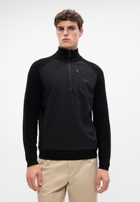Črna zip-up juha z visokim ovratnikom, ki se ponaša z teksturiranim materialom na straneh in detajlom logotipa na prsih. Gladko telo, prilegajoča se silhueta.