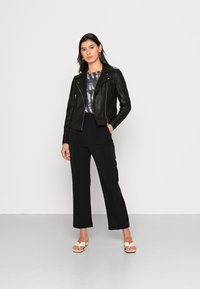 Jaqueta de motociclista em couro preto com zíperes prateados, combinada com um top cropped com efeito tie-dye cinza e calças largas pretas, complementadas por sandálias brancas.