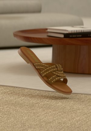 Sandale à enfiler marron cloutée avec des lanières croisées flottant au-dessus d'un tapis beige texturé, table basse ronde en bois et livres empilés en arrière-plan.