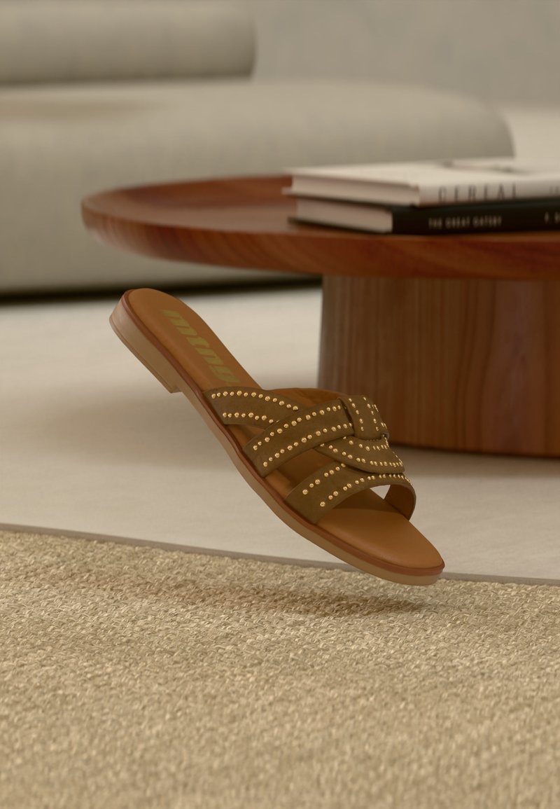 Sandale à enfiler marron cloutée avec des lanières croisées flottant au-dessus d'un tapis beige texturé, table basse ronde en bois et livres empilés en arrière-plan.