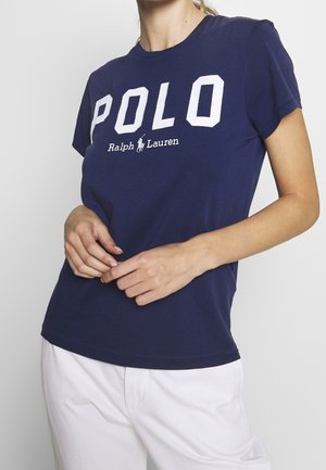 Femme portant un t-shirt polo Ralph Lauren bleu marine et un pantalon blanc, les mains légèrement jointes devant sa taille.
