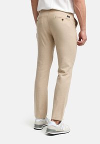 Pantalones de lino beige con corte recto, bolsillos traseros con cierres de botón, usados con zapatillas grises y blancas.