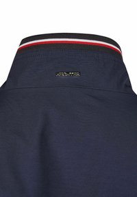 Marineblaue Jacke mit Kragen aus strukturiertem Stoff, gestreiftem Rippbündchen in Rot und Weiß und einer silbernen Metallschilder.