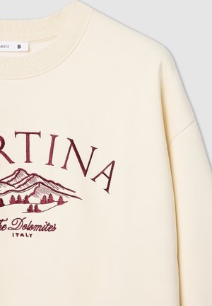 Kremowy sweter z żeberkowym, okrągłym dekoltem. Ma burgundowy haft z tekstem oraz wzór górski, wyróżniający się gładką, miękką strukturą.