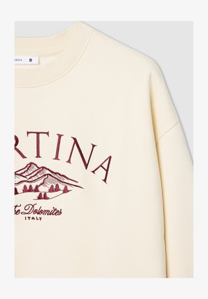 Kremowy sweter z żeberkowym, okrągłym dekoltem. Ma burgundowy haft z tekstem oraz wzór górski, wyróżniający się gładką, miękką strukturą.