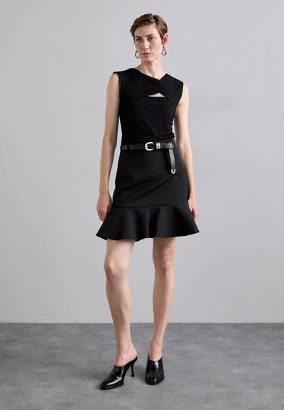 Haut sans manches noir avec détail en torsion, associé à une jupe noire ajustée avec un ourlet évasé et des mules à talons noirs brillants. Design minimaliste.