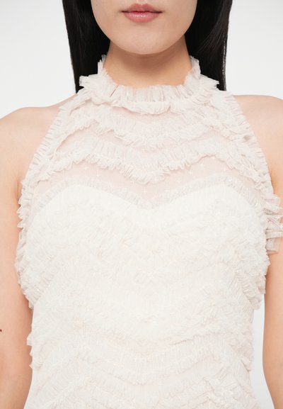 Biely volánový top na krk so skladanou textúrou látky a strapcami. Priehľadné detaily dodávajú hĺbku a zvýrazňujú dizajn výstrihu.