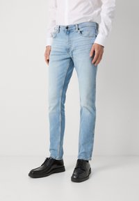 Světle modré džínové kalhoty s úzkým střihem, standardními kapsami a jemným vyblednutím. Nošeny s černými šněrovacími botami a bílou košilí.