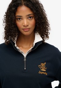 Marine blauwe zip-up sweatshirt met een witte kraag en een gouden geborduurd logo op de borst. Het heeft een zachte textuur en een zilveren rits.
