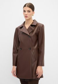 Bruine nepleer jas met een dubbelgebreid ontwerp, brede revers en twee voorzakken. Gladde textuur en zwarte knopenaccenten.
