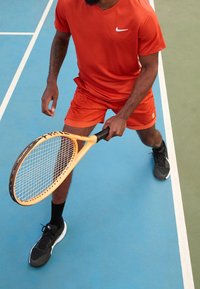 Oranžové sportovní tričko a šortky, černé sportovní boty, držící žlutou tenisovou raketu na modrém kurtu s bílými čarami.