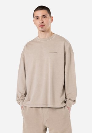 Beige hosszú ujjú ing puha anyagból, kerek nyakkivágással és kis logó részlettel a mellrészen. Laza szabás és egyszerű dizájn.
