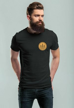 Schwarzes Baumwoll-T-Shirt mit einem runden, goldenen Baum-Graphic auf der linken Brust. Glatte Textur und klassischer Schnitt. Trägt es mit dunklen Jeans.