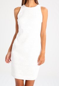 Vestido blanco sin mangas con una tela texturizada que presenta un patrón geométrico de triángulos. Diseño entallado, de longitud hasta la rodilla y con un escote redondo.