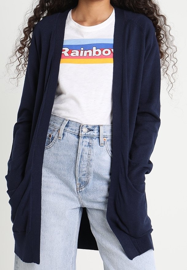 Marinblå cardigan med öppen framsida, långa ärmar och sidofickor, bärs över en ljus grafisk t-shirt och hög midja blå jeans.