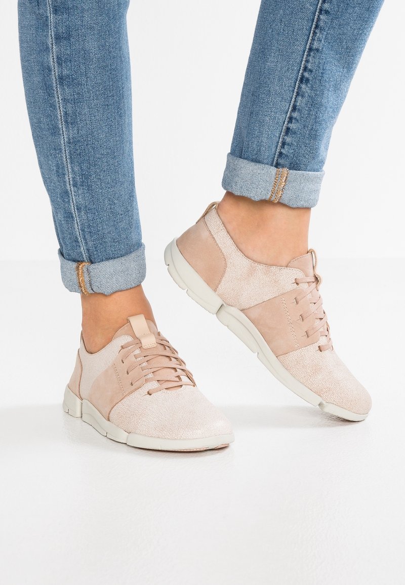 Pieds portant un jean bleu retroussé et des baskets décontractées rose clair avec des semelles blanches sur un sol blanc.