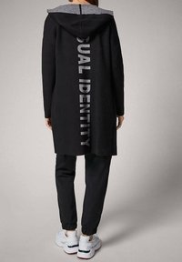 Svart lång hoodie med texten "DUAL IDENTITY" på ryggen, grå huva, mjukt tyg, kombinerad med svarta joggers och vita sneakers.