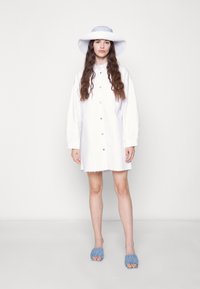 Vestuário branco com botões e mangas longas, com uma bainha desfiada; combinado com um chapéu de abas largas azul e branco com riscas e sandálias azuis franzidas.