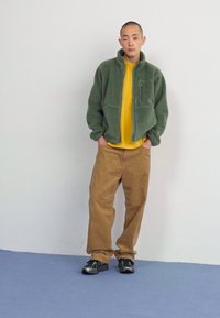 Zeleni flisani dukserica sa patentnim zatvaračem, žuta majica ispod, smešne kord pantalone i crne cipele. Model stoji protiv neutralne pozadine.