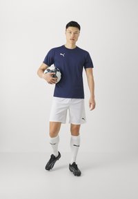 Marineblå kortermet skjorte, hvite shorts, sorte fotballsko, holder en fotball. Har PUMA-logo på skjorten og shortsene.