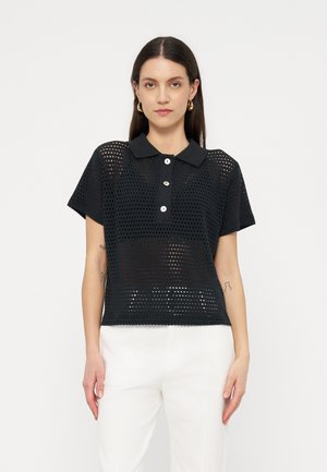 Hosszú sötét hajú nő fekete, rövid ujjú, hálós anyagú, fehér gombos poloingben, fehér nadrággal, egyszínű háttér előtt áll.