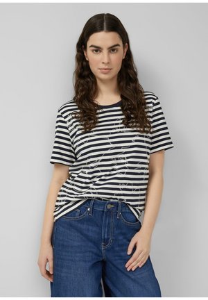 Kobieta z długimi, kręconymi, brązowymi włosami, ubrana w czarno-białą koszulkę w paski i niebieskie dżinsy, stojąca na tle jednolitego szarego tła.