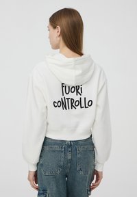 Felpa corta bianca con cappuccio e polsini elasticizzati, con la scritta nera "FUORI CONTROLLO" stampata sulla schiena. Tessuto morbido.