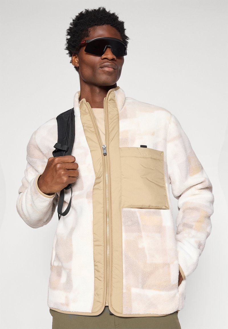 Fleecejakke i creme med beige detaljer, lynlåslukning foran og sidelommer. Har en kontrasterende tan-lommelap, glat tekstur.