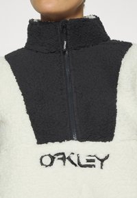 Schwarzer und weißer Fleece-Pullover mit einem Reißverschluss am Hals, ausgestattet mit einer strukturierten, einfarbigen schwarzen Schulterpartie und einem gestickten "OAKLEY"-Logo auf der Vorderseite.