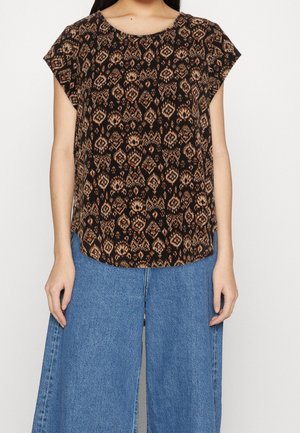 Femme portant un chemisier à manches courtes noir et marron à motifs, associé à un jean denim bleu à jambes larges.