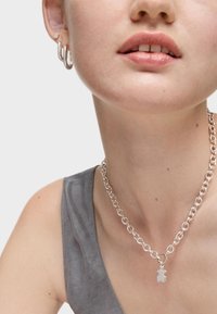 Collar de cadena de plata con un encantamiento de oso, llevado con pendientes de aro circulares. El modelo tiene piel clara y viste una blusa sin mangas gris.