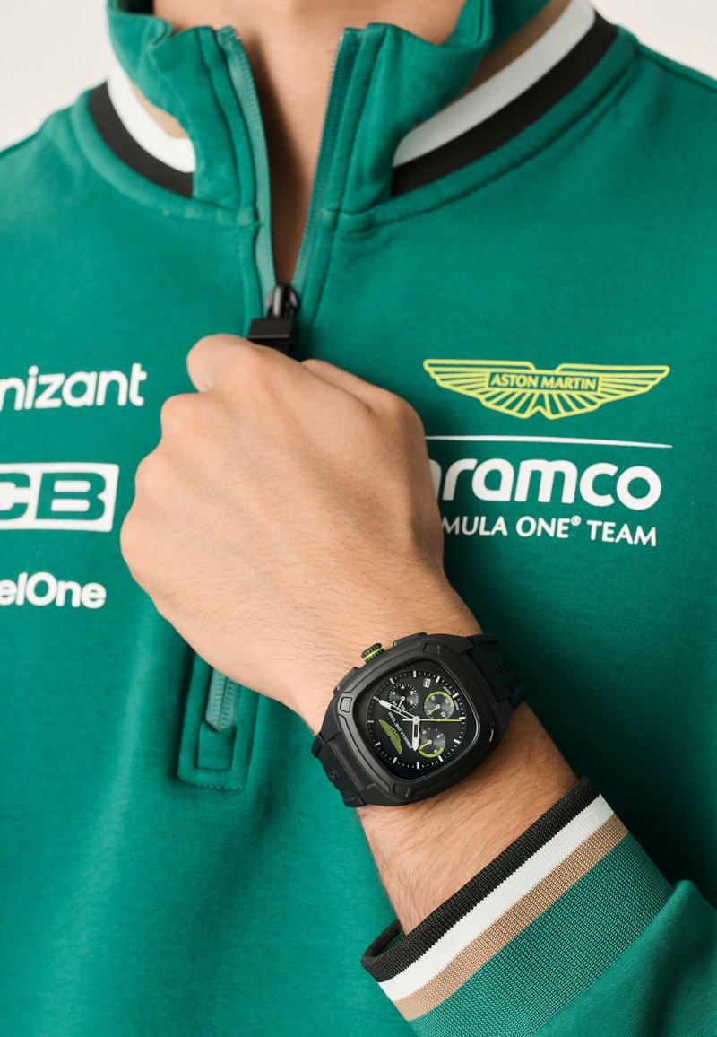 Melna sporta pulksteņa kvadrātveida ciparnīca ar zaļiem akcentiem un hronogrāfa funkcijām, kas valkāta uz rokas kopā ar tirkīzzilu džemperi ar rāvējslēdzēju un svītrainām aprocēm.