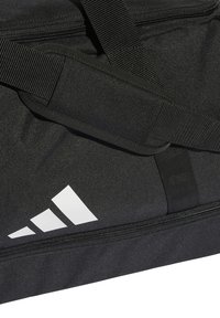 Crni torba s teksturiranim materijalom, s istaknutim bijelim Adidas logotipom i podesivim remenjem. Patentni zatvarač duž vrha.