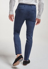 Homme marchant vu de dos, portant un pantalon slim à carreaux bleus, une chemise blanche et des mocassins en daim noirs sur un fond clair.