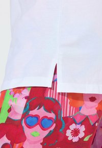Ourlet de chemise blanche avec fente latérale sur un tissu coloré présentant des visages stylisés, dont un avec des lunettes de soleil bleues en forme de cœur et du rouge à lèvres vert.