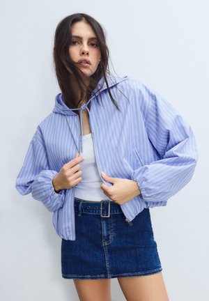 Junge Frau trägt eine hellblaue gestreifte Jacke mit Reißverschluss über einem weißen Oberteil und einem Jeans-Minirock und steht vor einem einfarbigen Hintergrund.