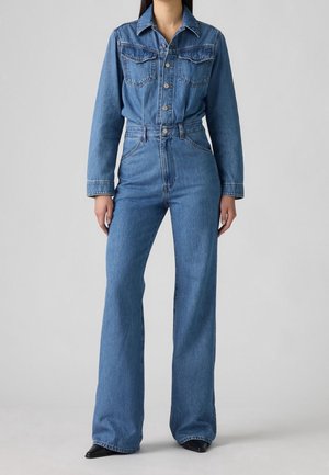 Femme portant une combinaison en denim bleu à manches longues, boutons sur le devant, poches poitrine et pantalon à jambes larges, debout devant un fond uni.