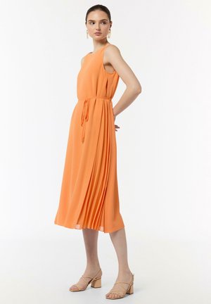 Orangenes ärmelloses Kleid mit einem Zugband in der Taille, gefaltetes Design, das unter das Knie fällt. Kombiniert mit hellen Blockabsatzsandalen.