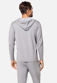 Uomo che indossa una felpa con cappuccio grigia chiara e pantaloni della tuta abbinati, in piedi con la schiena rivolta verso la camera su uno sfondo neutro.