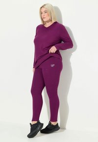 Kvinna med blont hår som bär en matchande lila långärmad tröja och leggings med svarta sneakers, stående mot en vit vägg.