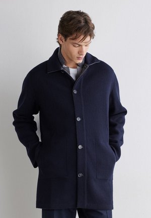 Junger Mann mit braunen Haaren trägt einen dunkelblauen, geknöpften Mantel mit den Händen in den Taschen und blickt nach unten vor einem einfarbigen hellgrauen Hintergrund.